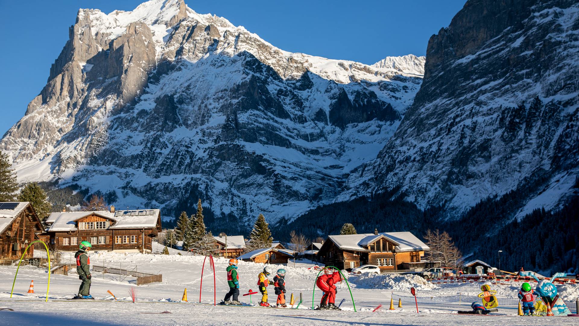 Children with skis and helmets on a ski slope, accompanied by an adult, in front of snow-covered mountains.